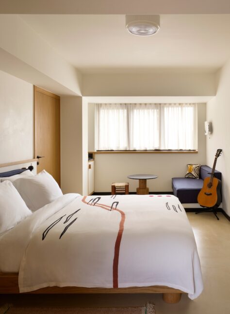 Cozy hotel room with a soft, white duvet and abstract design. A guitar leans against a blue sofa near a window. Warm tones, serene atmosphere.