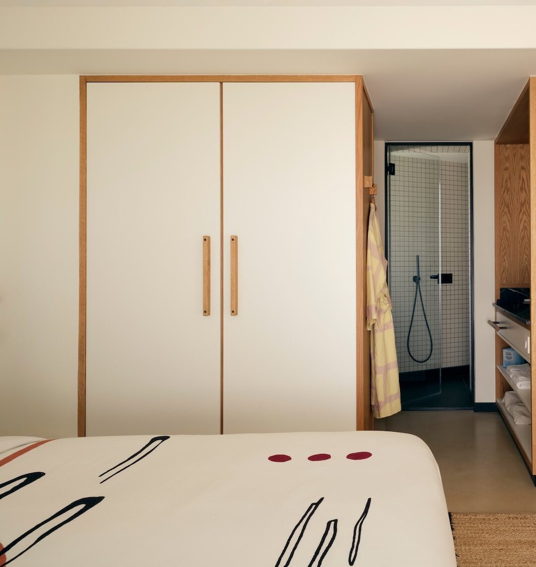 Cozy hotel room with a minimalist design. Features a bed with abstract-patterned bedding, a large wardrobe, open shelving, and a glimpse of a modern bathroom.