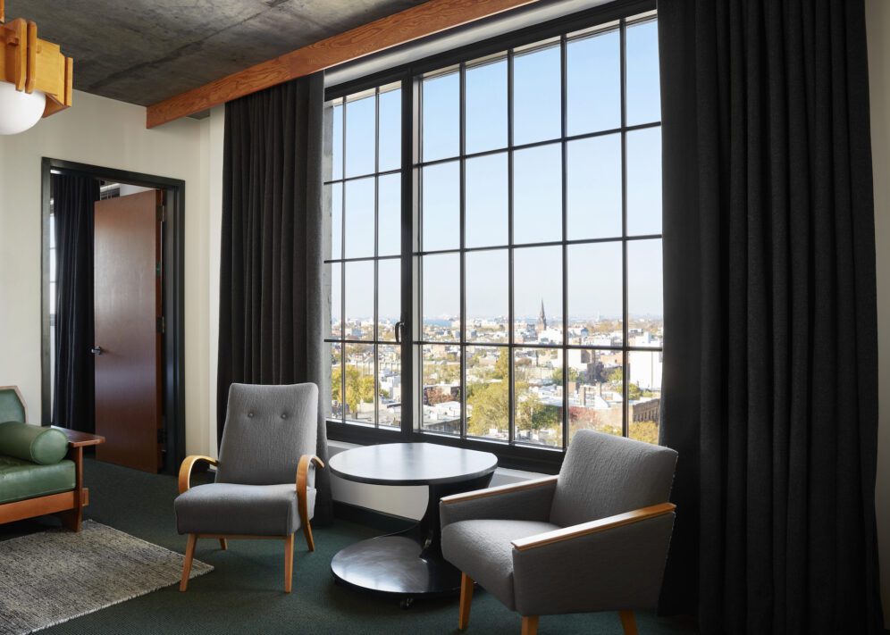 Two gray armchairs and a round table are positioned by a large window with black curtains, overlooking a cityscape.