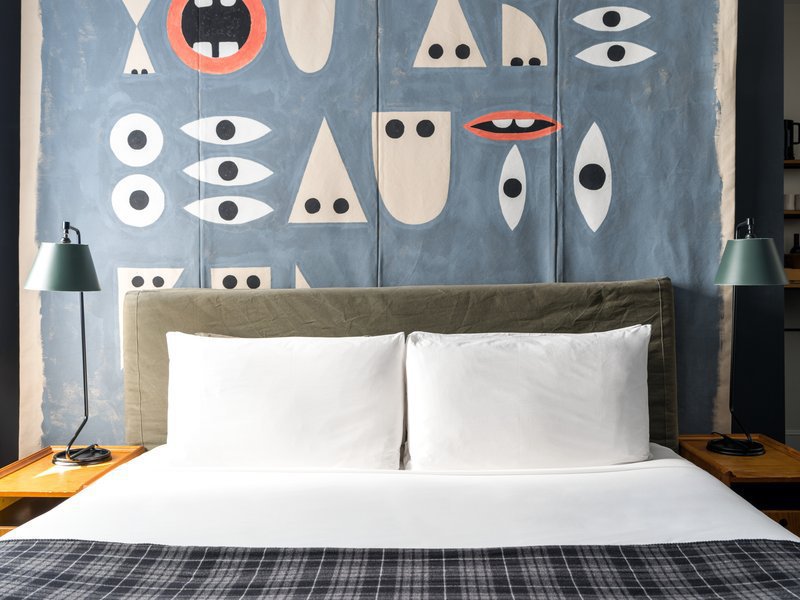 A bed with plaid covers is set against a wall featuring floor to ceiling custom art. Lamps on either side of the bed sit on small tables