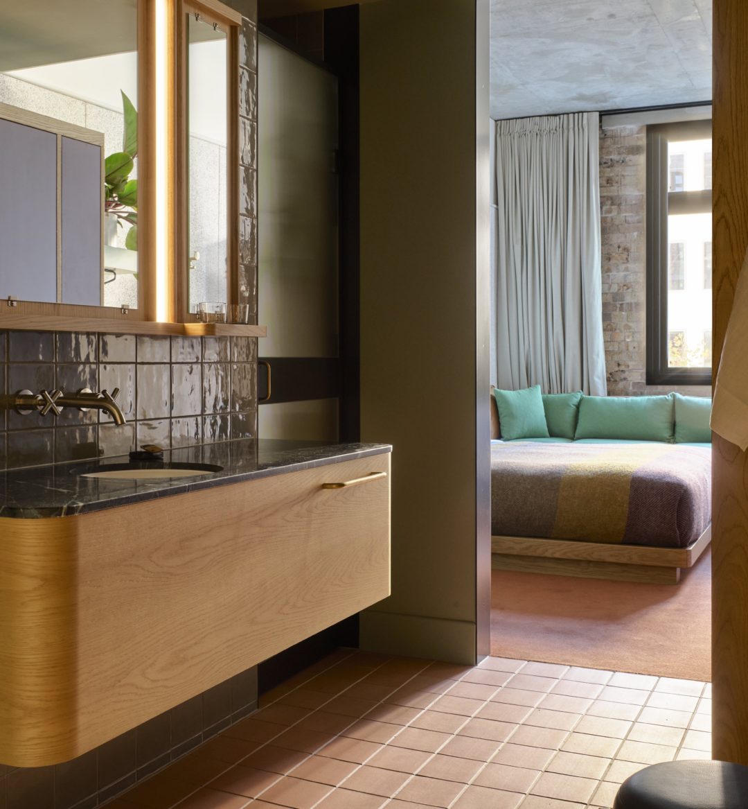 A sleek tiled wall supports to knobs that provide water to a black granite sink, and an entryway to the bed area is in the background