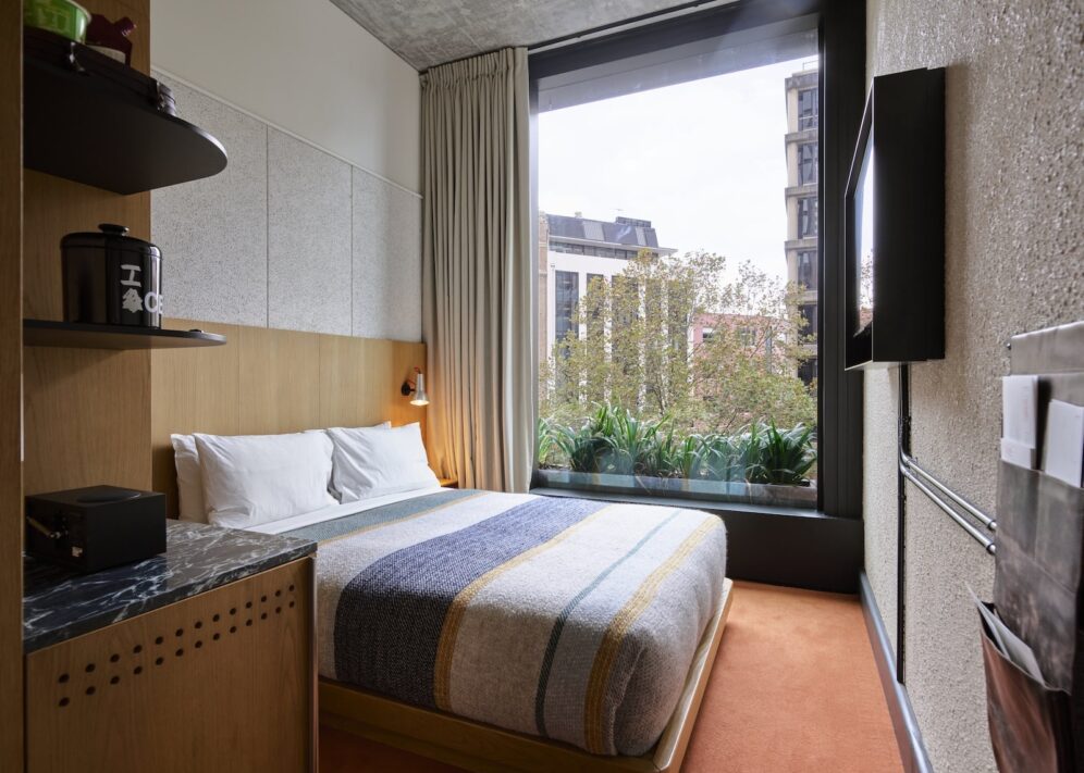 A cozy room features a bed with stylish linens and a wall length window looking over greenery outside