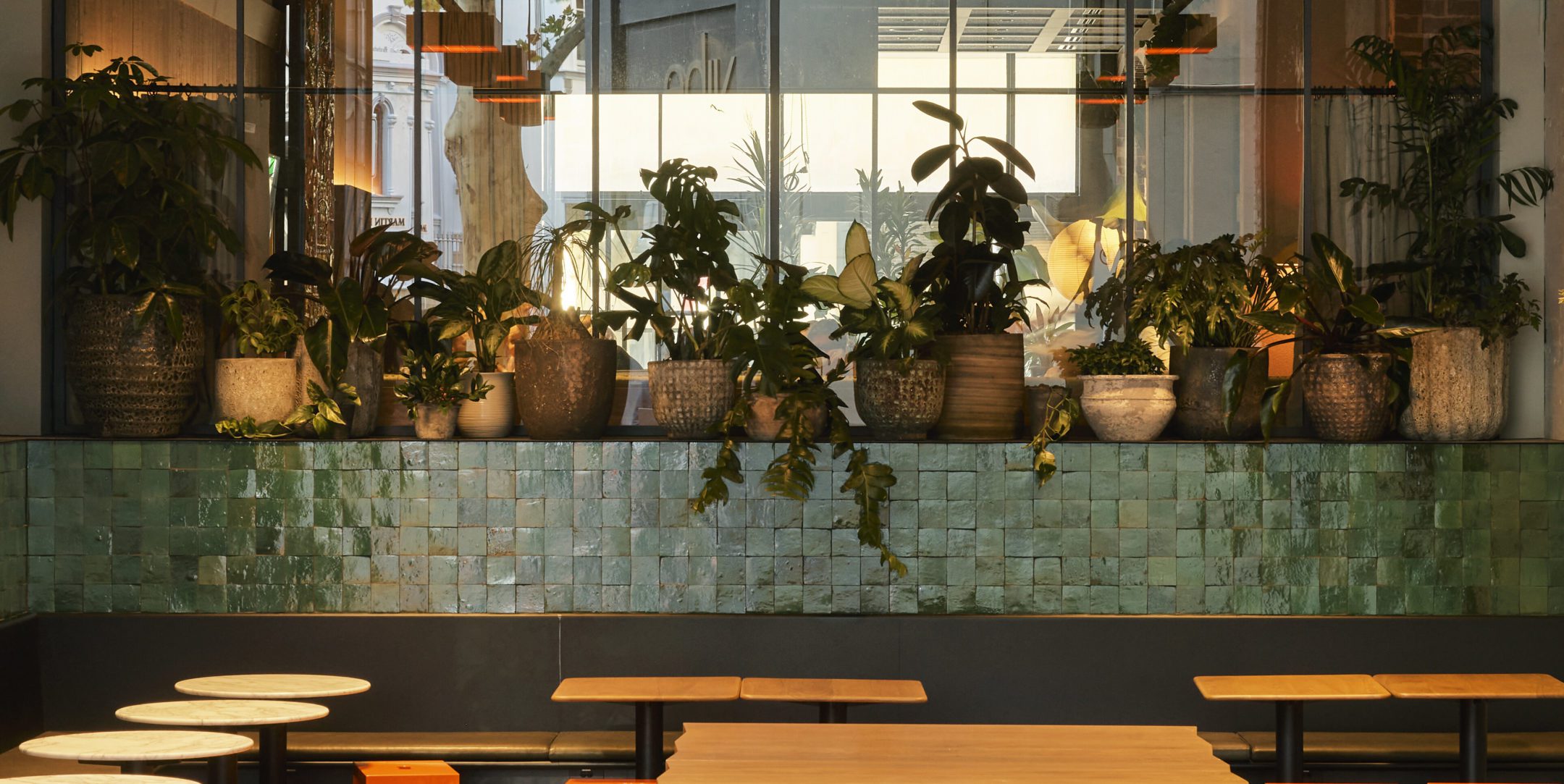 Indoor seating area with wooden tables and green tiled wall, decorated with potted plants arranged on a ledge in front of a mirror.
