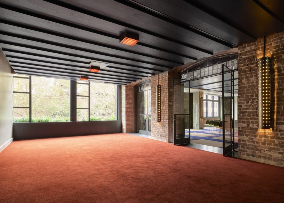 An event space with exposed brick walls, orange/red carpet and a wall length window providing views to the outdoors