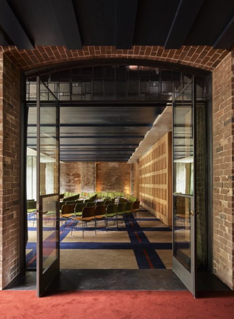 Glass doors open to a modern conference room with rows of green chairs, exposed brick walls, and patterned carpet.