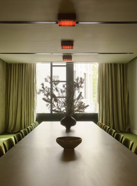 Minimalist dining room with a long table, green chairs, green carpeting, large window, and a vase with branches as centerpiece.