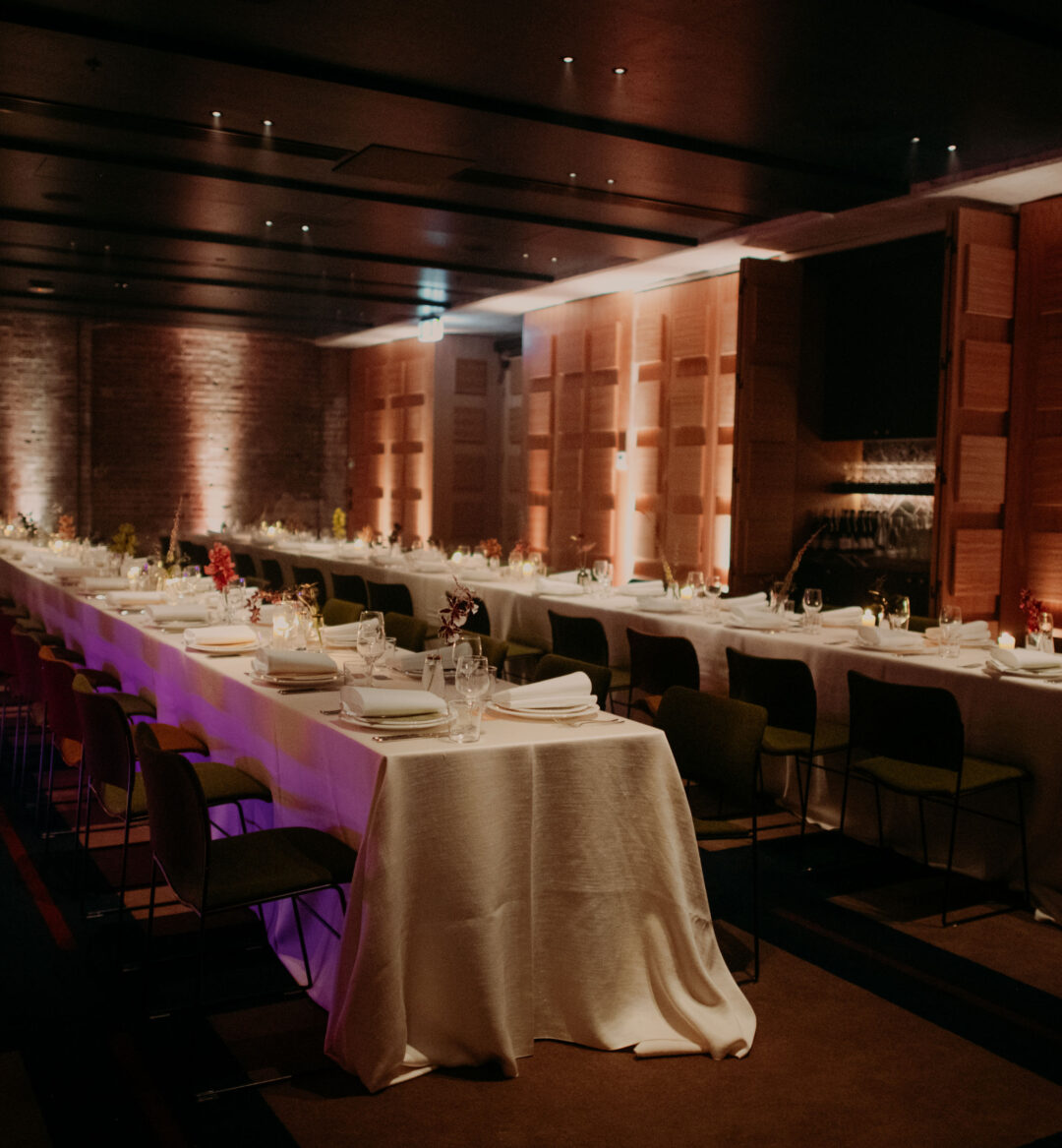 Two long banquet tables set for a formal dinner in a dimly lit, modern event space with brick and wood-paneled walls.