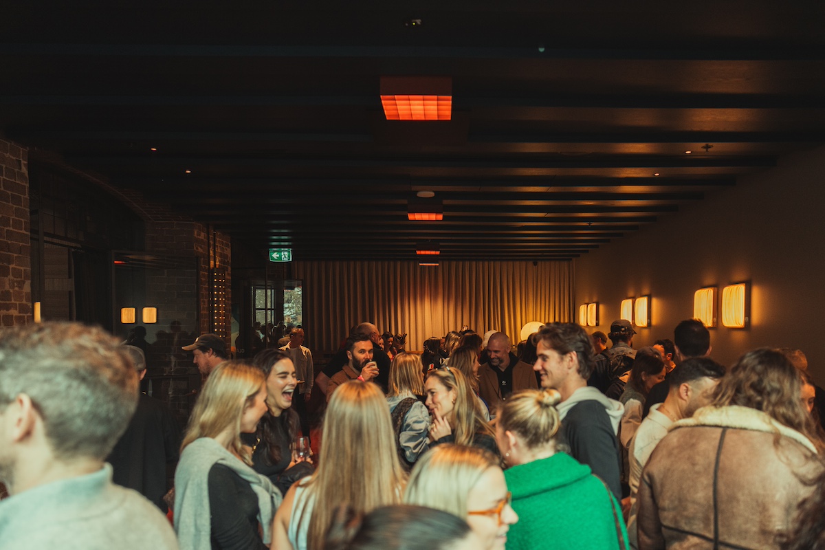 A large group of people socializing in a dimly lit indoor space with wooden beams and warm wall lights.