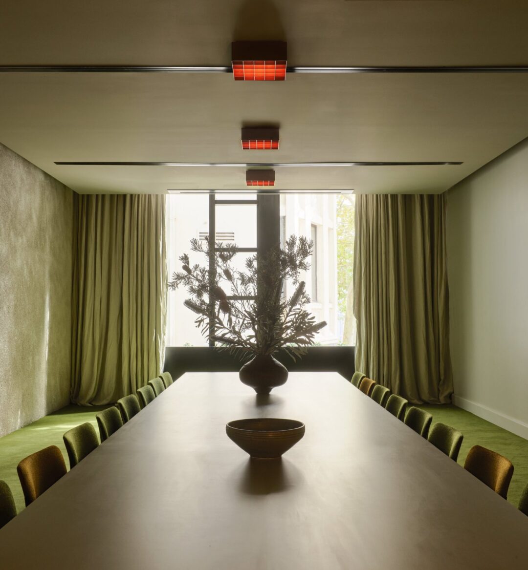 Minimalist dining room with a long table, green chairs, green carpeting, large window, and a vase with branches as centerpiece.