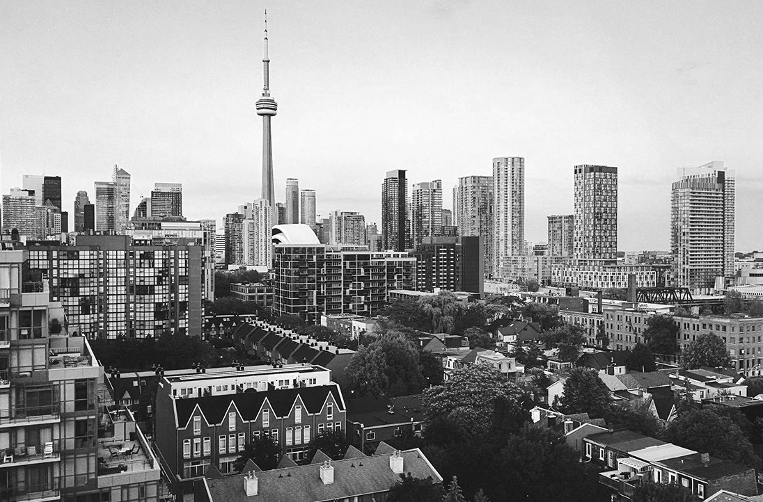 Black and white photo of Toronto’s skyline with the CN Tower and high-rise buildings above residential houses and trees.