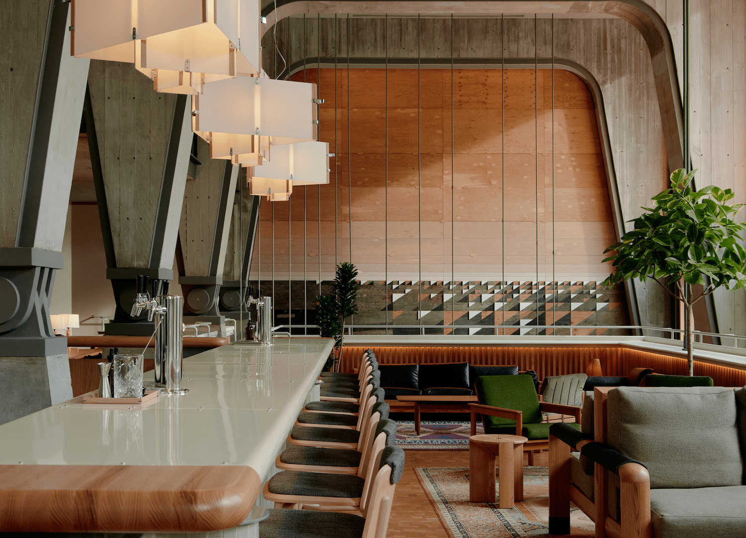Modern cafe interior with a long bar, wooden chairs, pendant lights, and green lounge seating under large arched concrete beams.